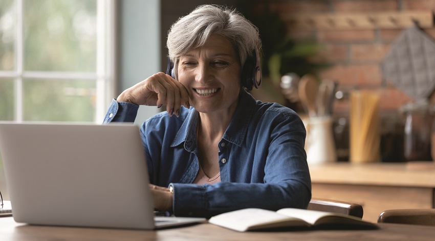 An older woman smiles while watching a computer screen with her notebook open beside her