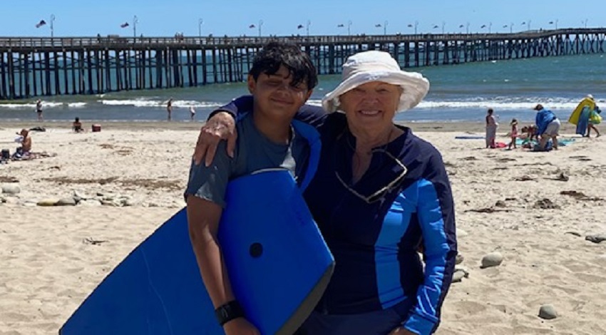 Grandparent and grandchild on the beach in front of a boardwalk, ready to go boogie boarding.